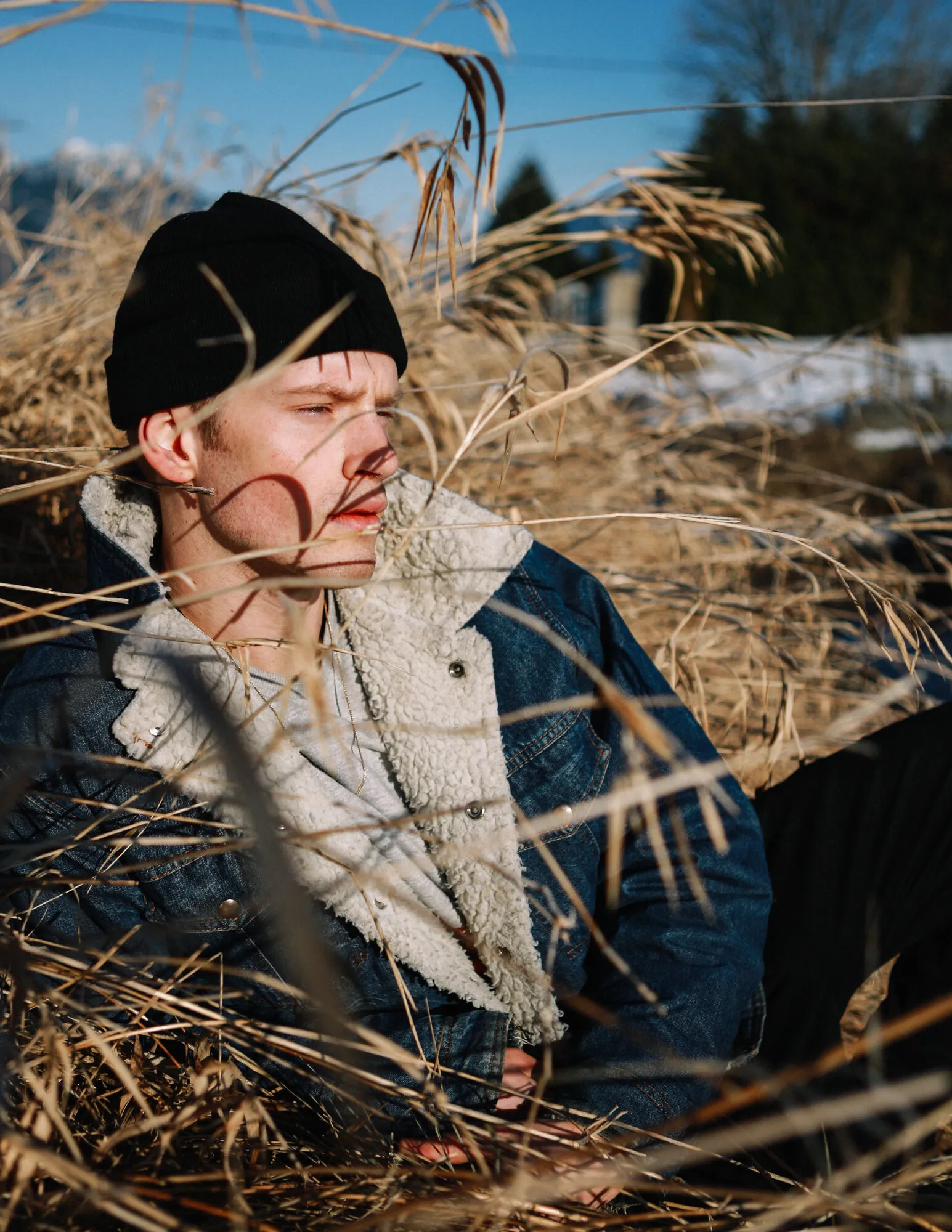 Model in a black beanie crouched among tall dried grasses in denim