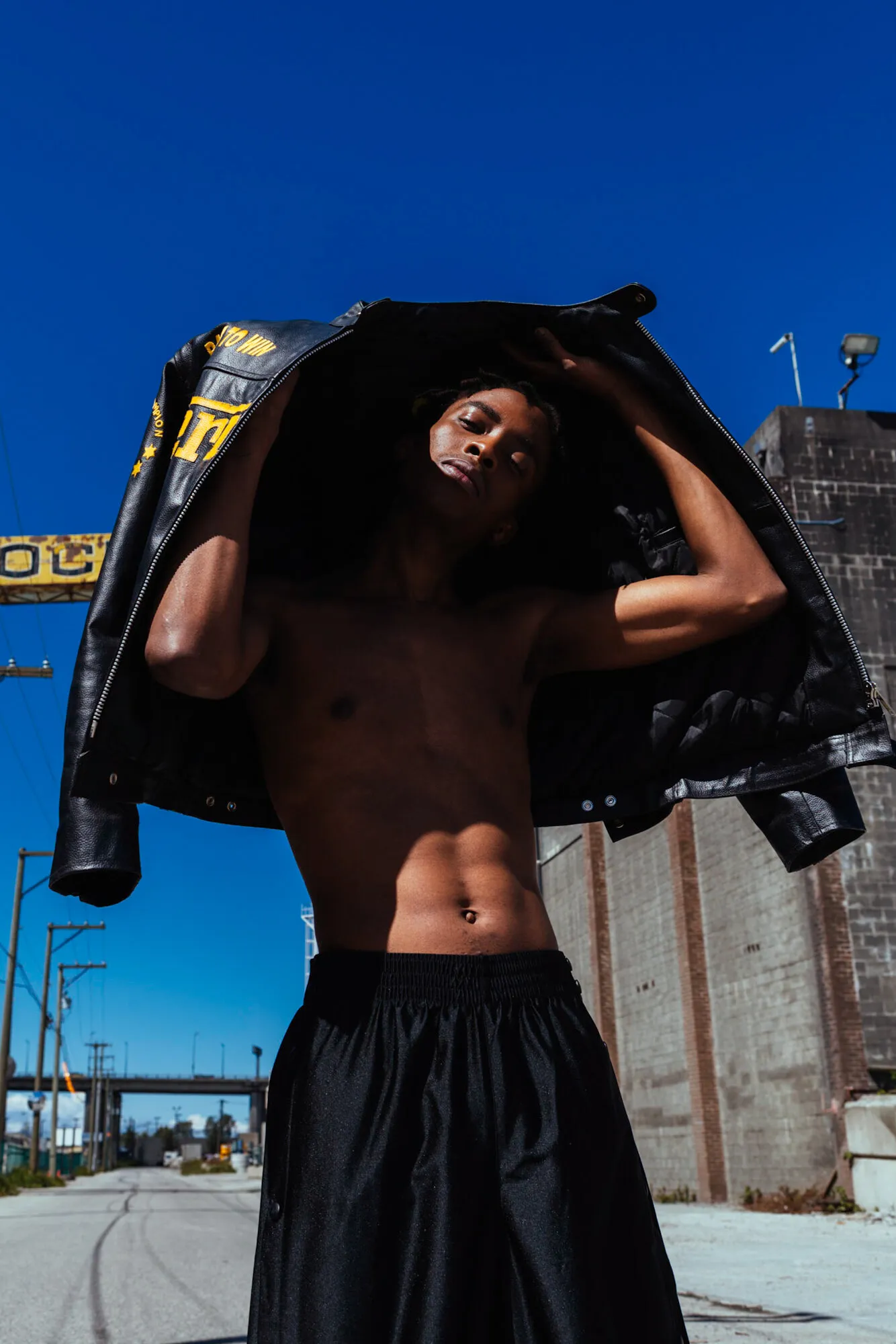 Model pulling a leather jacket overhead against a deep blue sky on an industrial street