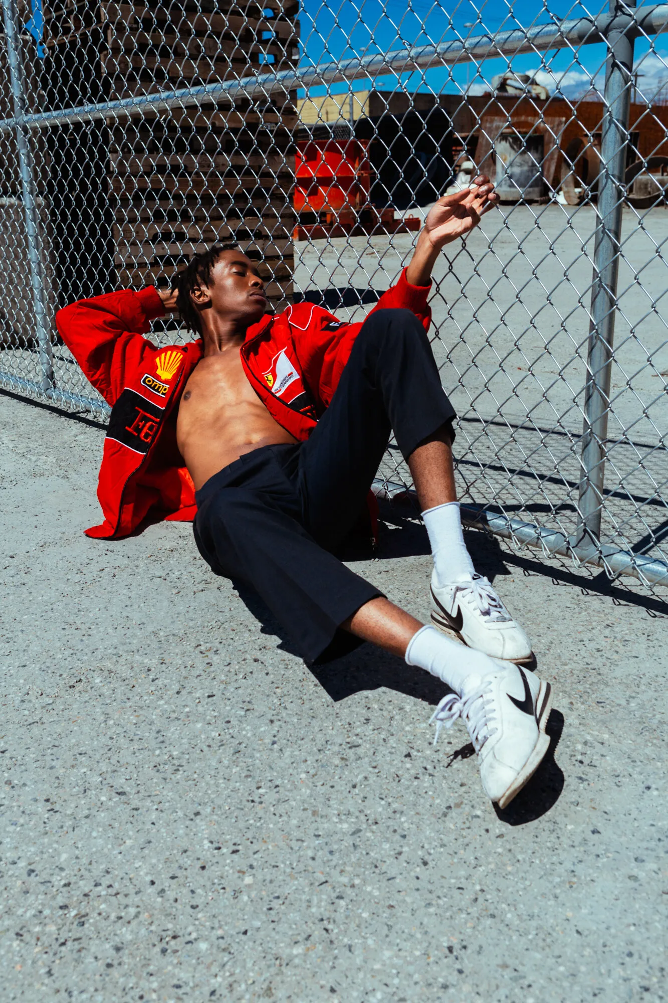 Model in a red jacket reclining on pavement beside a chain-link fence