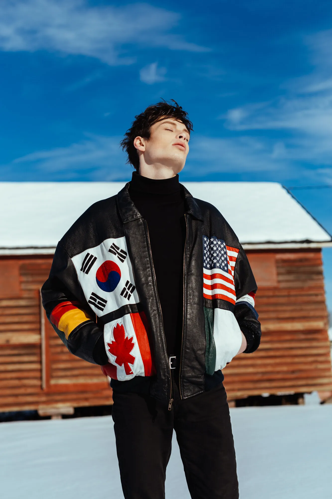 Model in a bomber jacket patched with international flags, standing before a snow-covered cabin