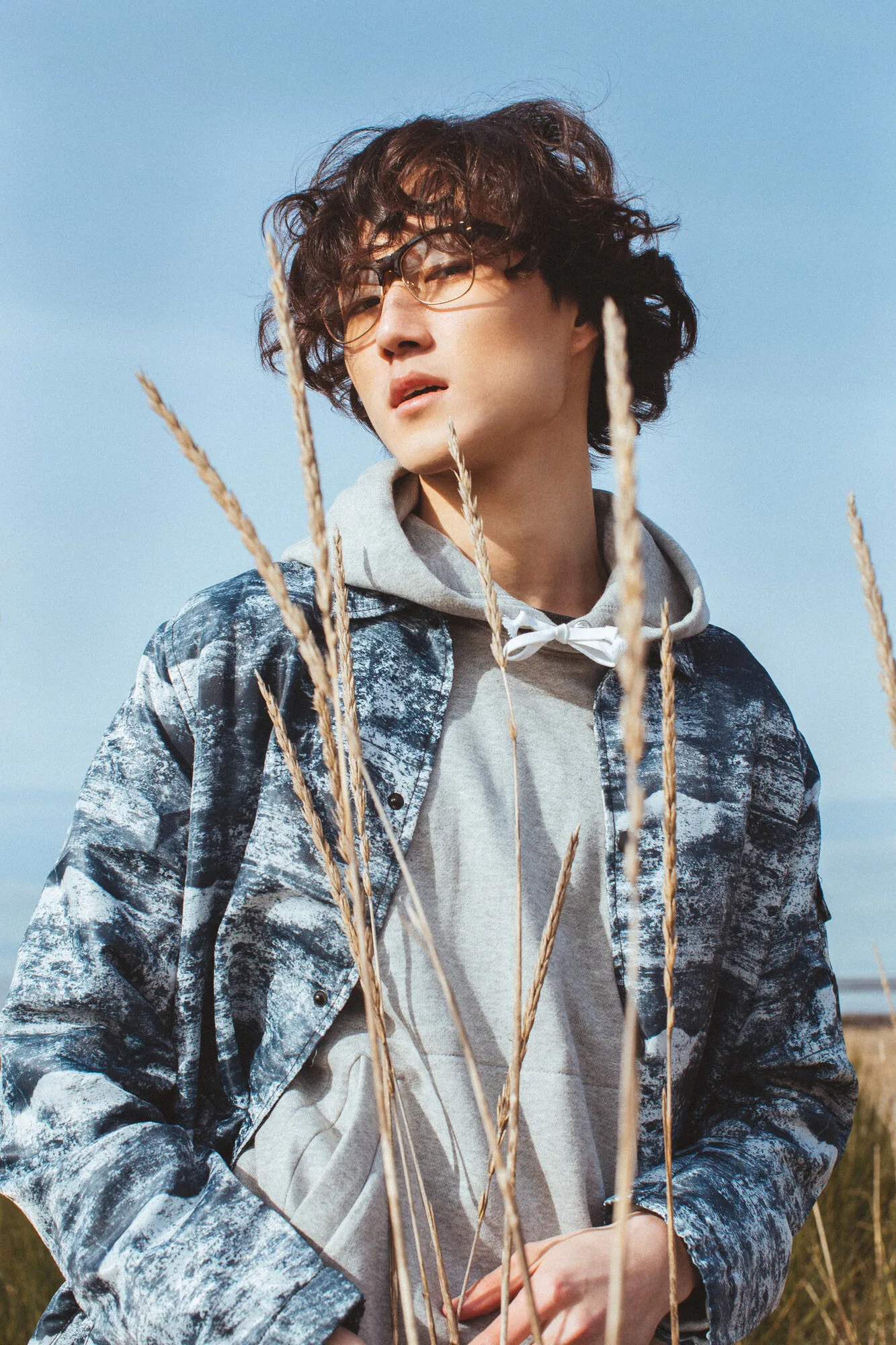Model in a patterned blue jacket and grey hoodie photographed against the sea and sky